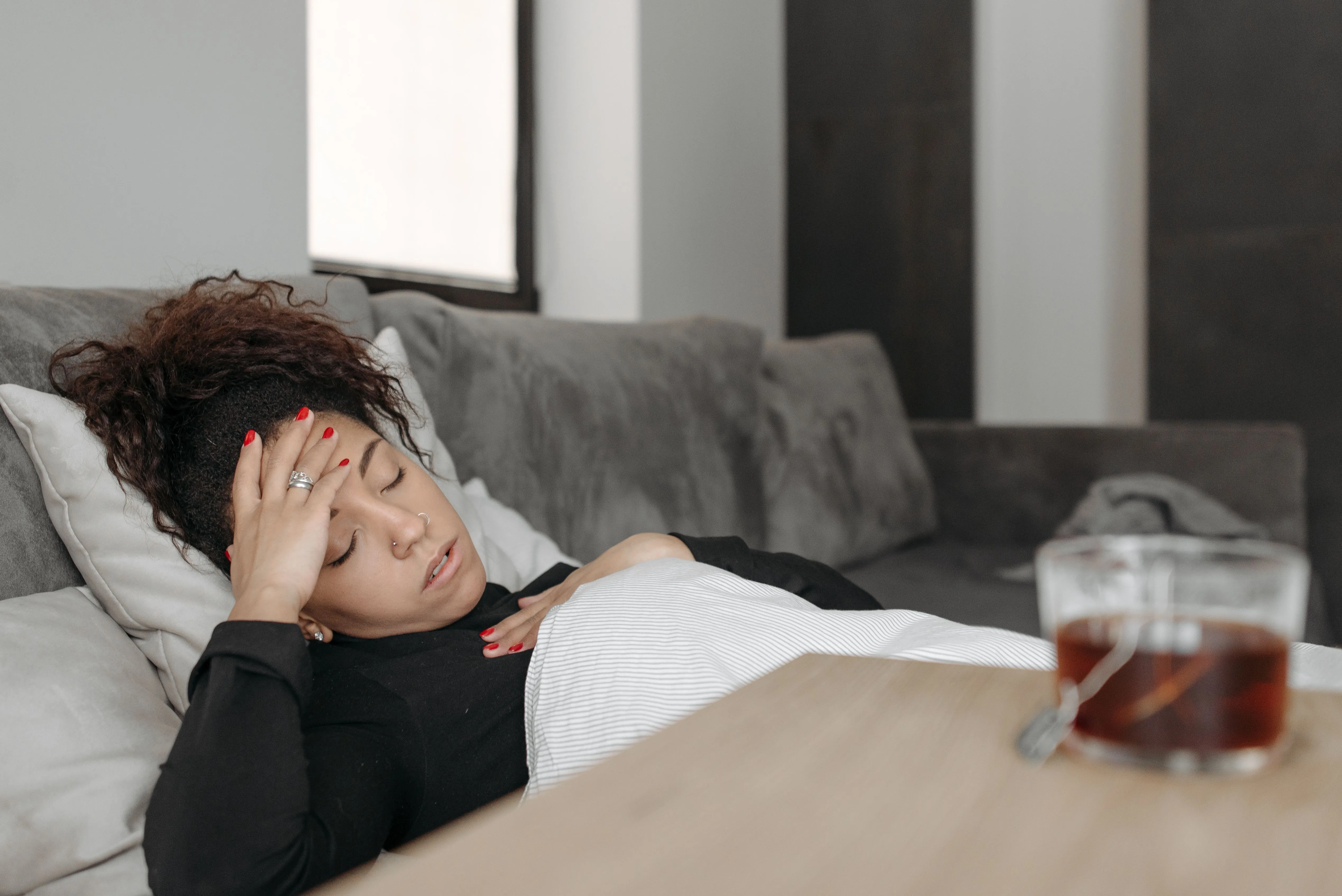 A woman is laying on a sofa feeling unwell, a cup of tea is next to her.